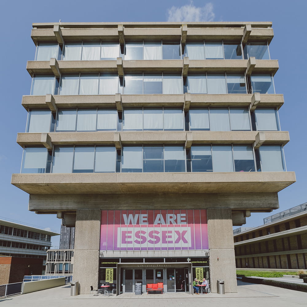 External view of the Albert Sloman Library. The photo is taken straight on from the front of the library, making the library appear deceptively square. There's four floors with large windows, the sun gleaming off them. Below the windows is signage that says 'We Are Essex', and on the ground floor there are doors leading into the Lakeside Theatre Café. 