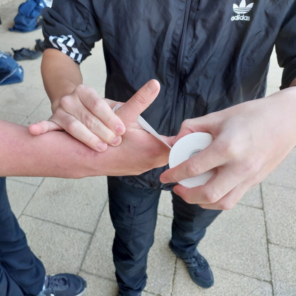 Two people, both mostly out of shot. The one on the left is having their hand wrapped up with tape by the person on the right.